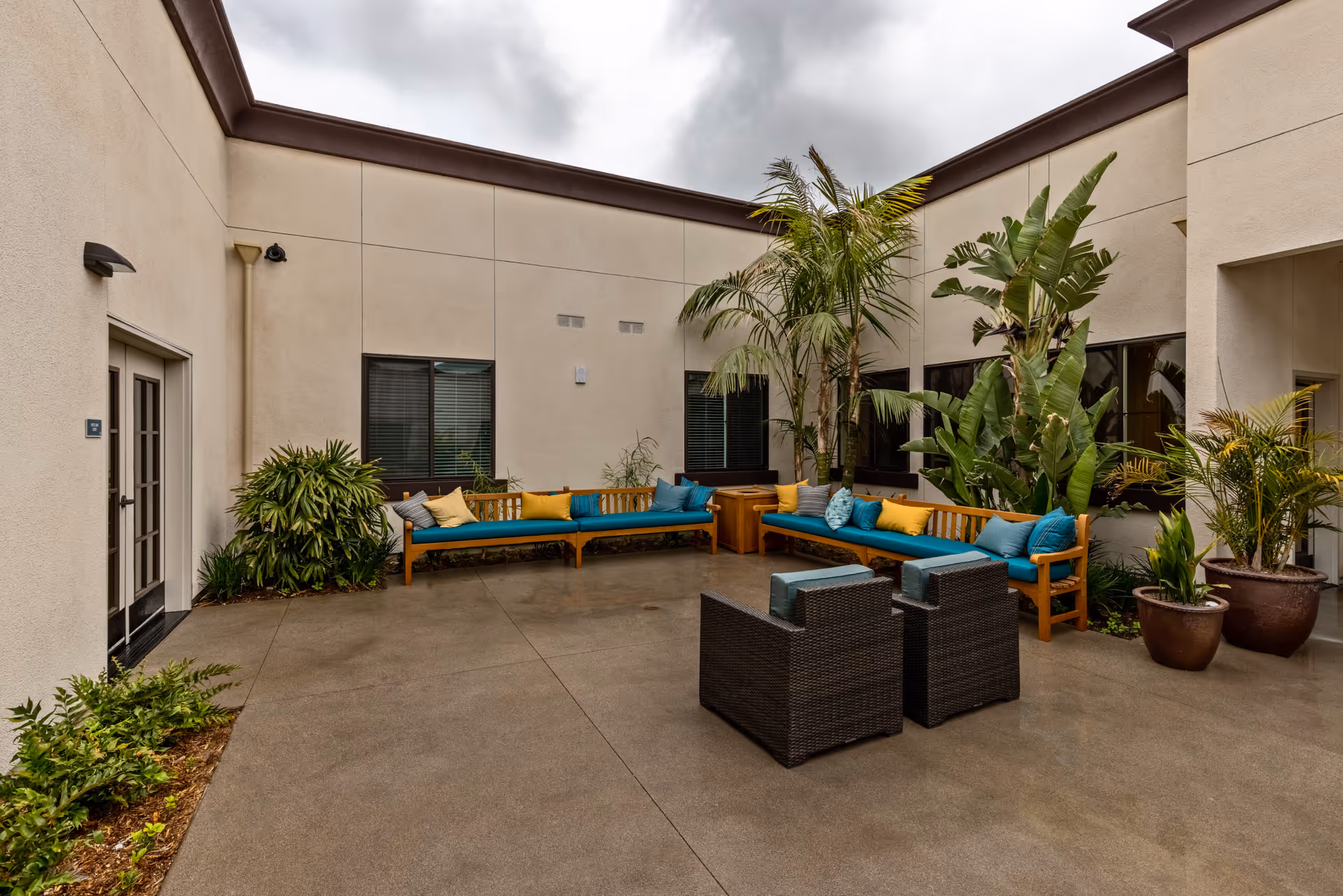 Outdoor courtyard area with beige walls, large windows, and a variety of green plants including palm trees. There are wooden benches with blue cushions and colorful throw pillows arranged in an L-shape, along with two dark wicker chairs with blue cushions in the center on a concrete floor under a cloudy sky.