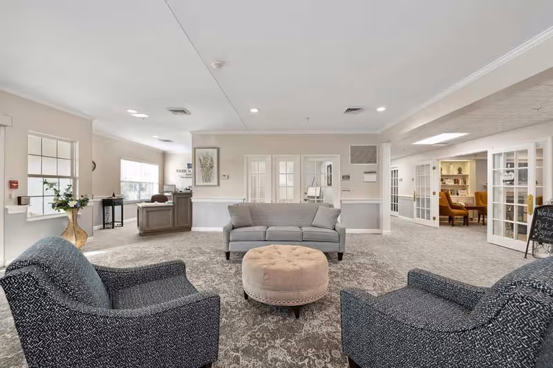 A spacious and well-lit senior living facility common area with a gray sofa, two patterned armchairs, and a round beige ottoman in the center. The room features light-colored walls, carpeted flooring, and multiple glass-paneled doors leading to other rooms. A reception desk is visible in the background with a sign that reads 'Park Square Manor'. There is also a small chalkboard sign near the entrance welcoming visitors.