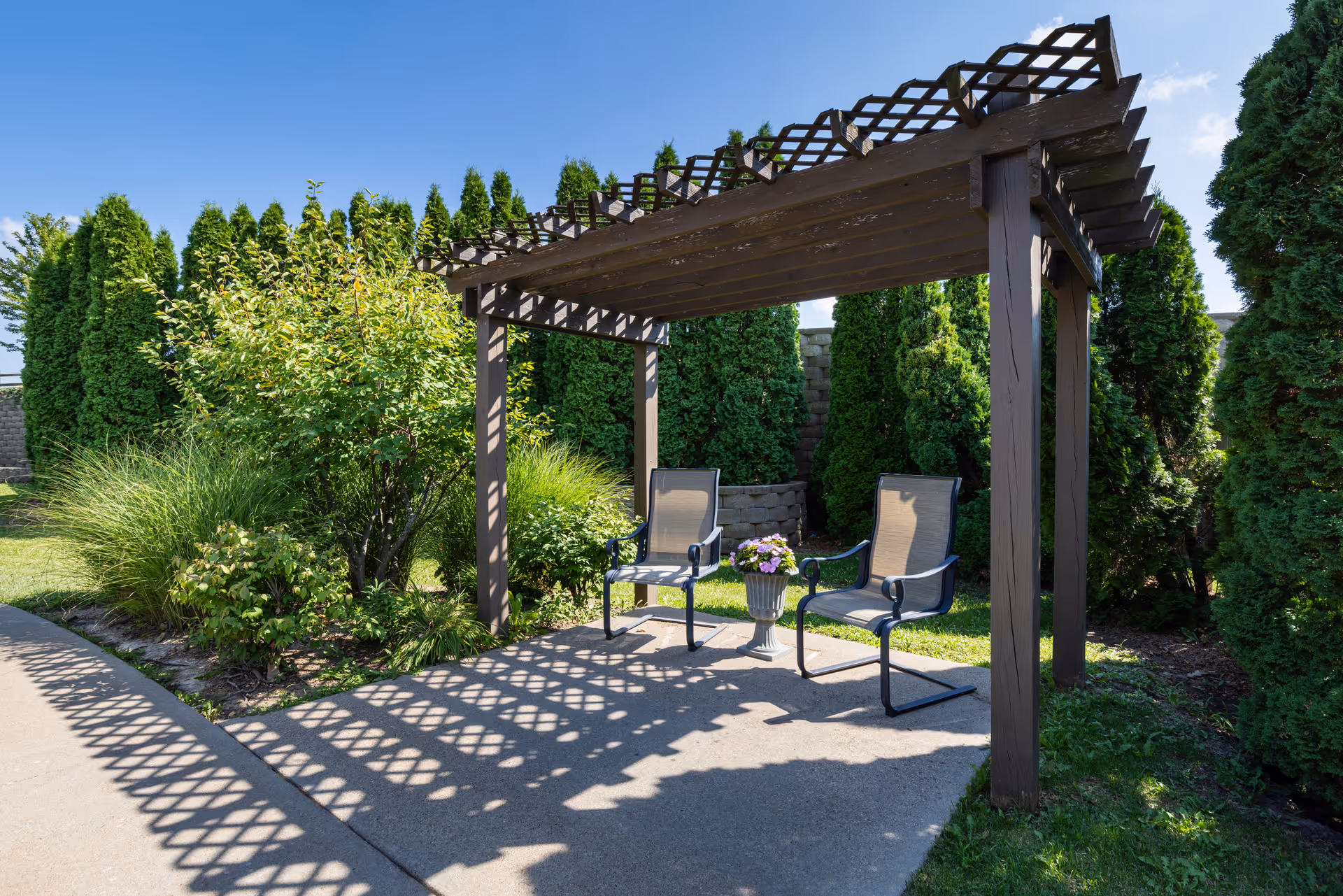 Two outdoor chairs and a planter under a wooden pergola in a landscaped garden area.
