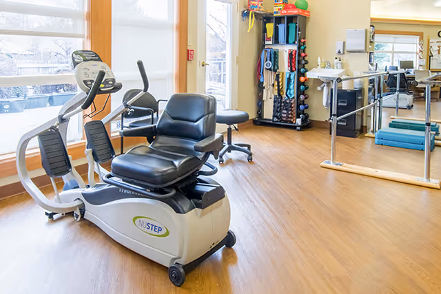 A bright exercise room with a NuStep recumbent cross trainer, a small stool, a rack with various exercise equipment, a ballet barre, and large windows letting in natural light.