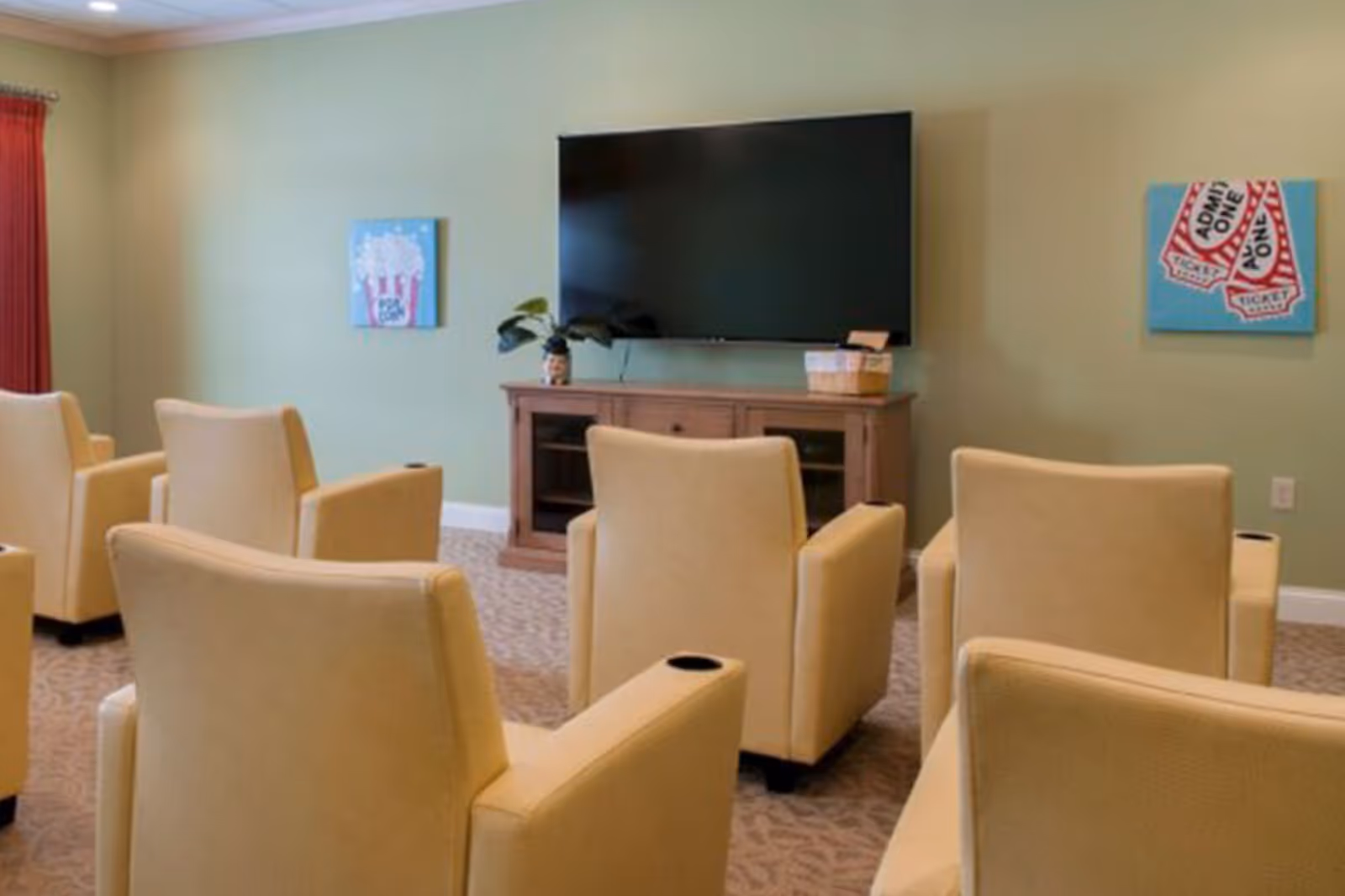 A small theater-style room with several beige armchairs arranged in rows facing a large flat-screen TV mounted on a light green wall. Below the TV is a wooden cabinet with a plant and a basket on top. The walls have two colorful paintings, one of popcorn and the other of movie tickets. A red curtain is partially visible on the left side.