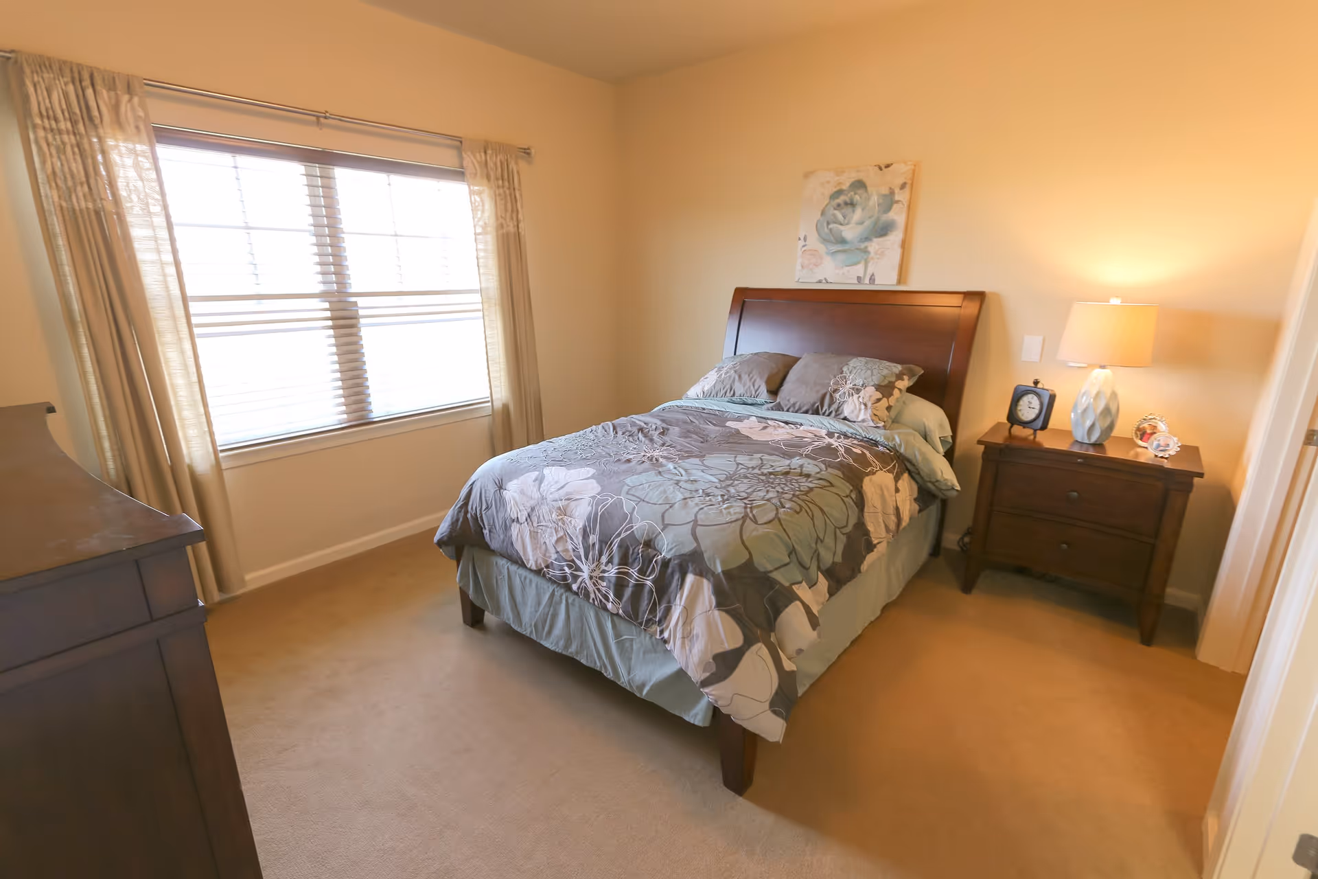 A cozy bedroom with a wooden bed frame and floral-patterned bedding. There is a window with beige curtains on the left wall, a wooden dresser partially visible on the left, and a wooden nightstand with a lamp, clock, and small decorative items on the right. A floral painting hangs on the wall above the bed.