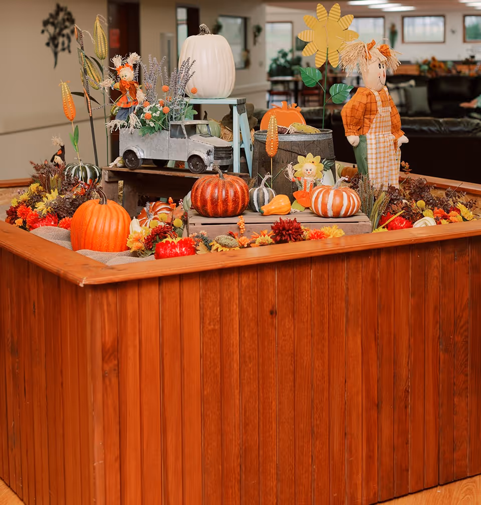 A wooden planter box filled with autumn decorations—pumpkins, flowers, a toy truck and a scarecrow—displayed in a communal indoor area.