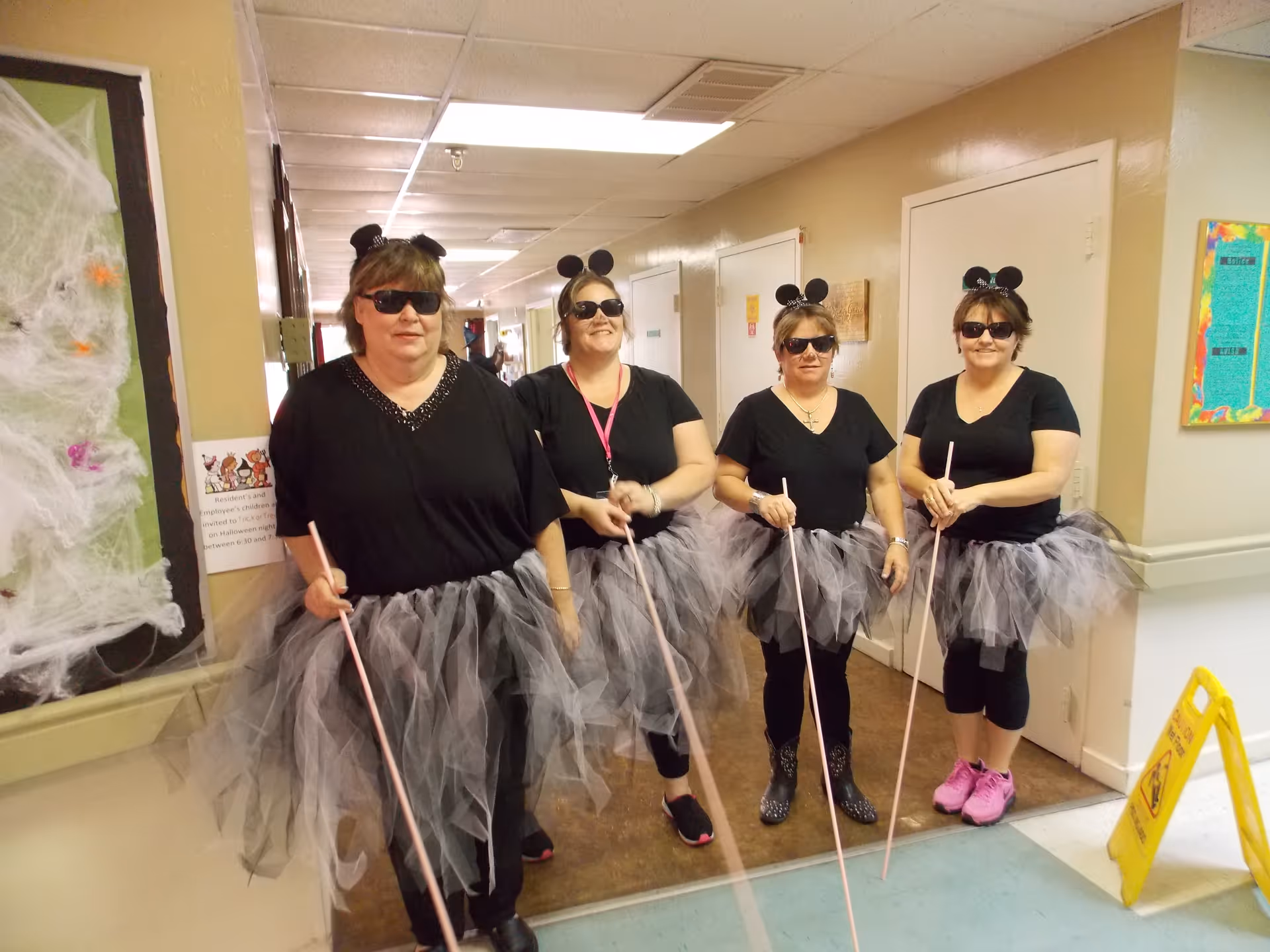 Four women dressed in black shirts, black and gray tutus, black mouse ears headbands, and sunglasses standing in a hallway holding white canes, possibly celebrating a themed event at a nursing and rehabilitation center.