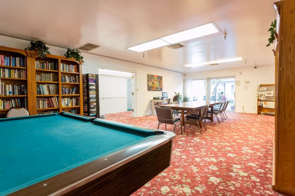 Interior room with a pool table in the foreground, a large wooden bookshelf filled with books, a table with chairs, and a red floral carpet. The room has white walls, fluorescent ceiling lights, and a glass door leading outside.