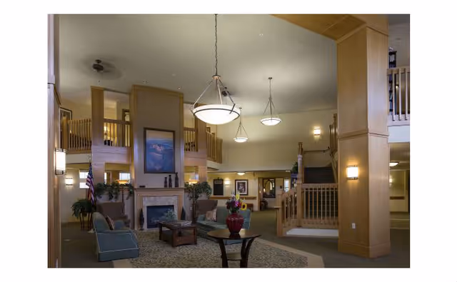 Spacious senior living facility common area with high ceilings, wooden pillars, a fireplace, comfortable seating including armchairs and sofas, decorative plants, and hanging light fixtures. A staircase leads to an upper balcony overlooking the room.