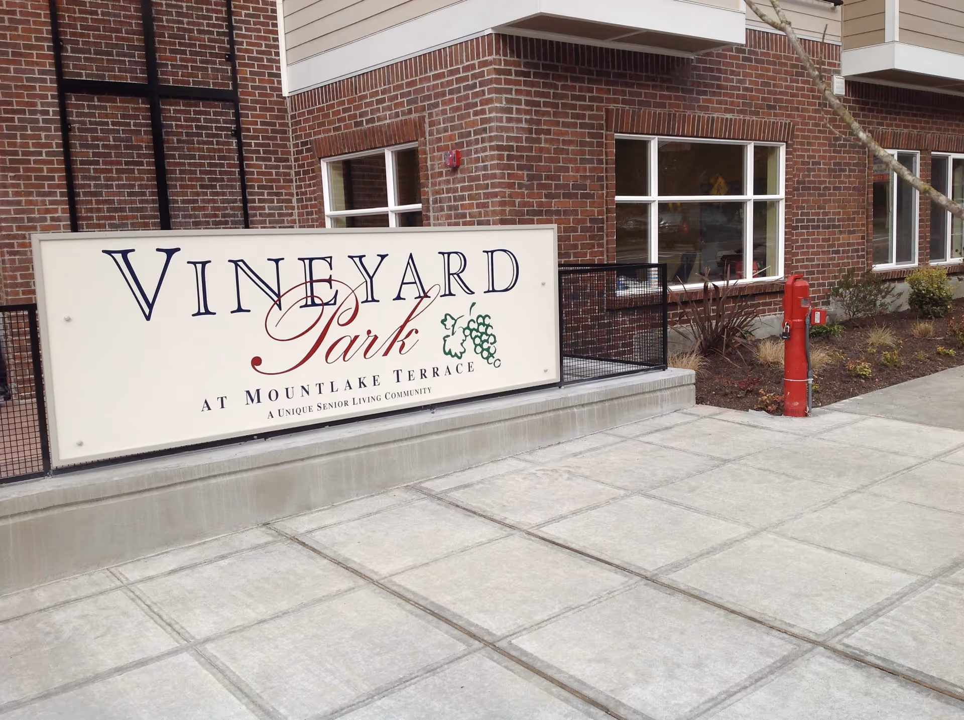 Outdoor view of the entrance area of Vineyard Park at Mountlake Terrace, showing a large sign with the facility name in front of a brick building with windows and some landscaping.