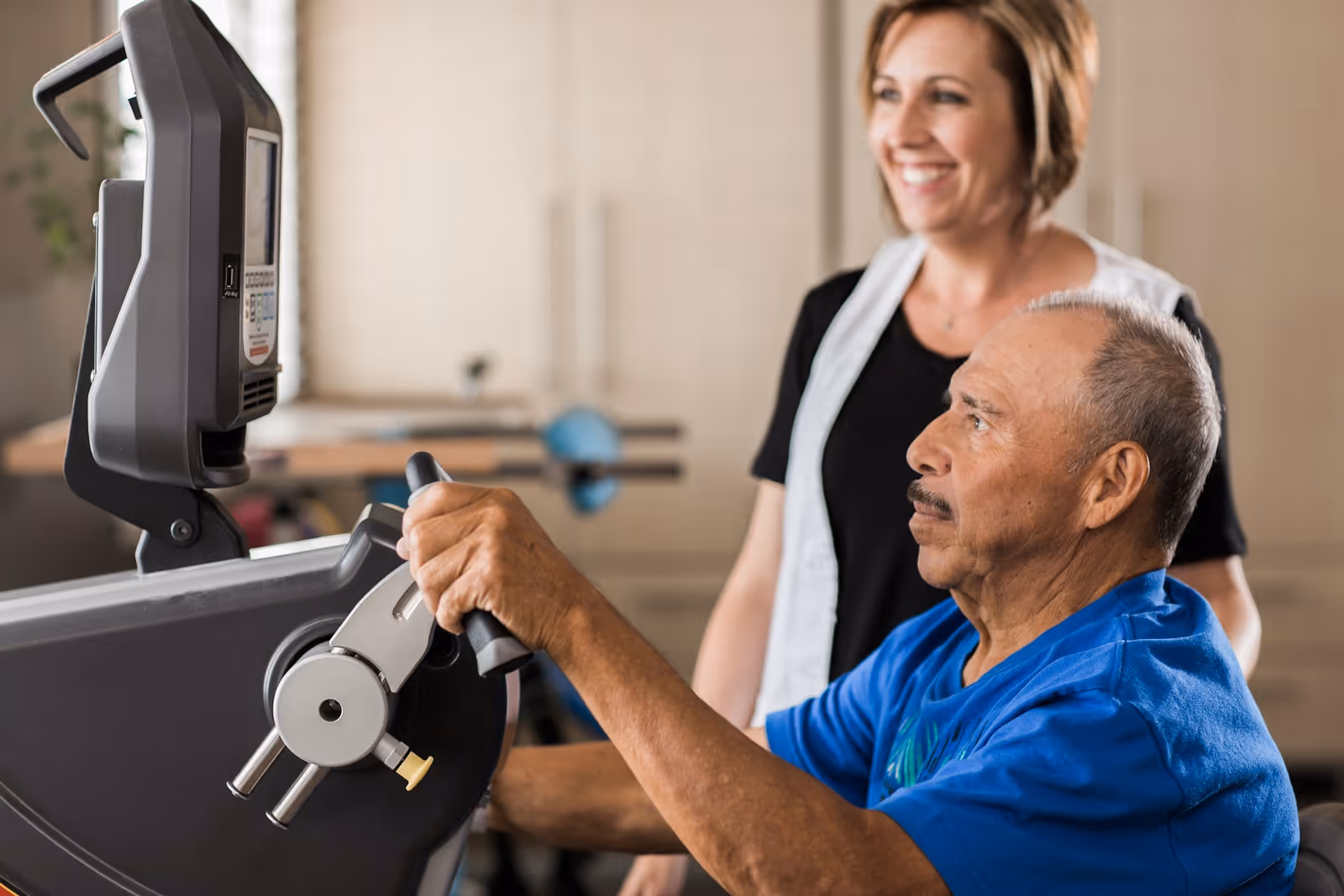 An elderly man in a blue shirt is using a piece of exercise equipment while a smiling female caregiver or physical therapist stands behind him, providing support and encouragement in a rehabilitation or therapy setting.