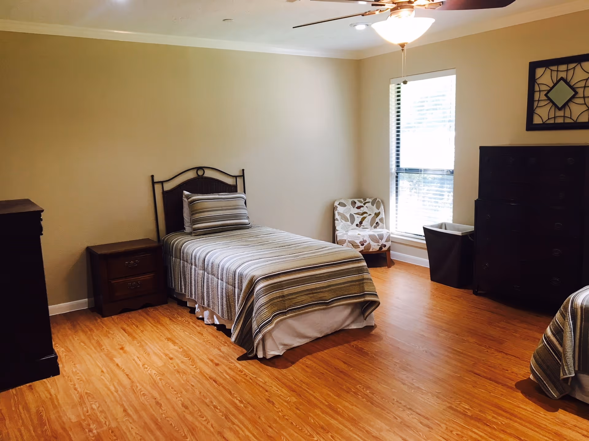 Light-filled bedroom with a single bed, nightstand, dresser, patterned chair, and hardwood floors.
