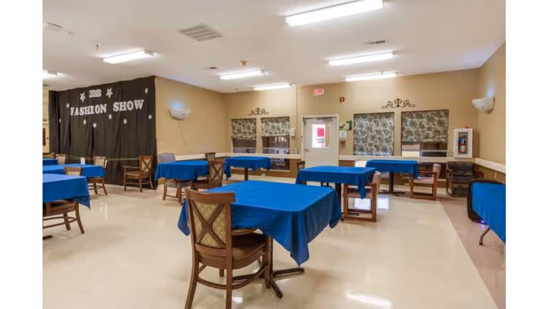 Community dining/activity room with multiple tables covered in blue tablecloths and wooden chairs and a curtained stage area labeled 'Fashion Show'.