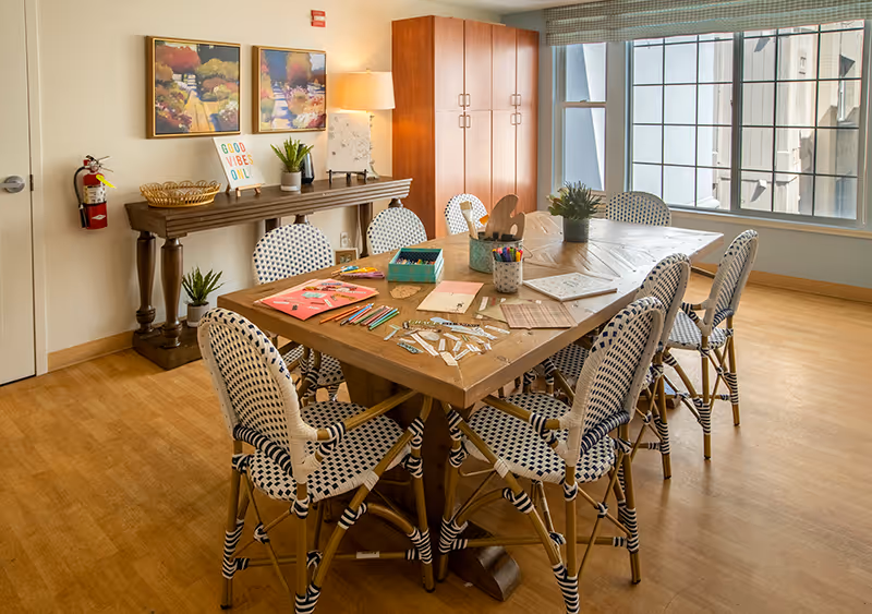 A well-lit room with a wooden table surrounded by eight patterned chairs. The table is covered with various art supplies including colored pencils, markers, paper, and craft materials. In the background, there is a wooden console table with decorative items and two colorful paintings on the wall. A large window lets in natural light, and a wooden cabinet stands against the wall.