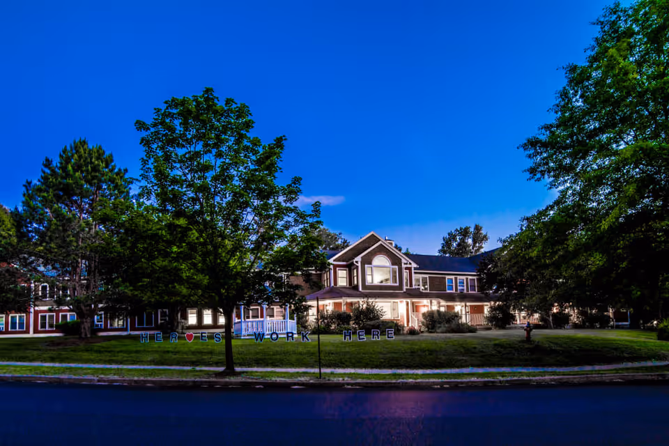Exterior view of a senior living facility building at dusk with lights on inside. The building is surrounded by trees and a well-maintained lawn. In front of the building, there is a sign on the grass that reads 'HEROES WORK HERE' with a heart symbol.