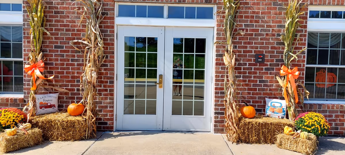 Double glass doors with white frames set in a red brick wall, decorated with autumn-themed items including hay bales, pumpkins, corn stalks tied with orange ribbons, potted yellow flowers, and decorative pillows with fall designs.