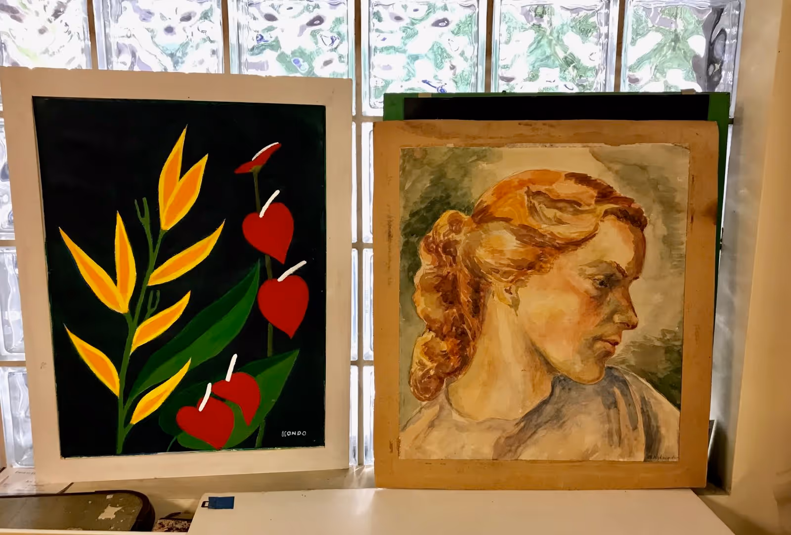 Two paintings displayed side by side in front of a glass block window. The left painting features stylized yellow and red flowers with green leaves on a black background. The right painting is a portrait of a woman with reddish hair styled in a braid, looking to the right, painted in warm tones.