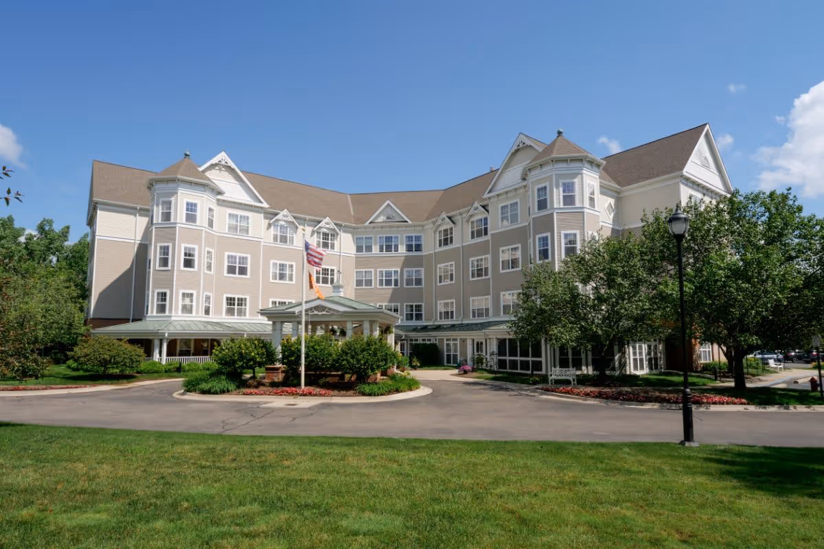 Exterior view of a large, multi-story assisted living facility building with beige siding and white trim, surrounded by green trees and a well-maintained lawn under a clear blue sky. There is a circular driveway with a flagpole displaying the American flag and another flag in front of the entrance.