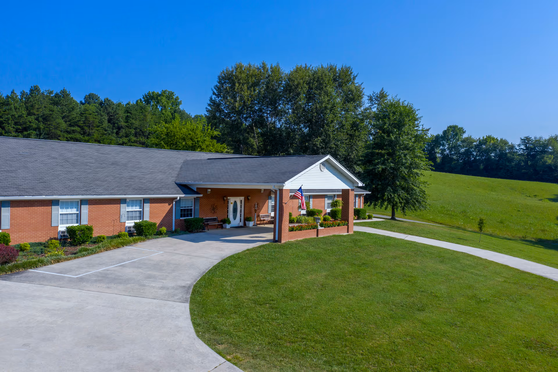Exterior view of a single-story brick building with a gray roof, white trim, and an American flag near the entrance. The building is surrounded by a well-maintained lawn, trees, and a curved concrete driveway under a clear blue sky.