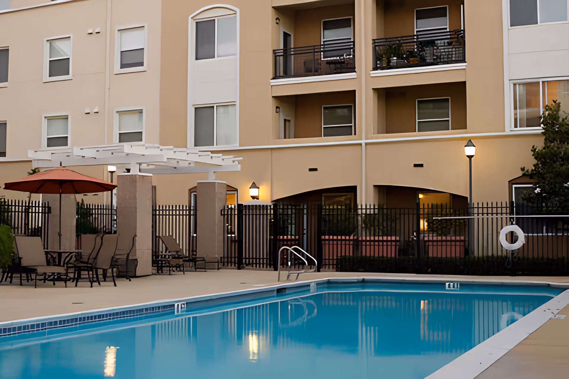Outdoor swimming pool area at Heritage Estate Senior Apartments with clear blue water, surrounded by a concrete deck. There are several lounge chairs and tables with umbrellas near the pool. The background shows a multi-story beige apartment building with balconies and windows, some with plants and patio furniture.