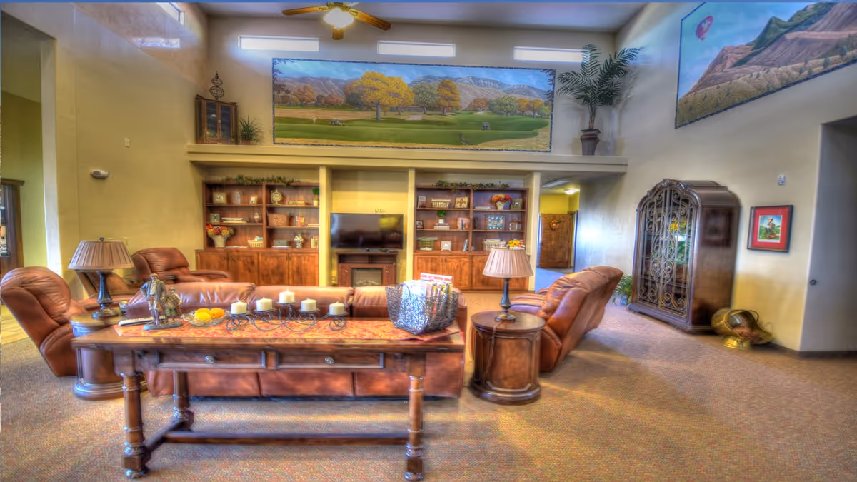 A spacious communal living room with leather sofas, wooden tables and shelves, a TV, and a large landscape painting on the wall.