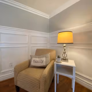 A cozy corner featuring a beige upholstered armchair with a decorative pillow that says 'family', next to a small white side table holding a modern table lamp with a beige shade and a small potted plant. The walls have white wainscoting with a light gray upper section and crown molding, and the floor is wooden.