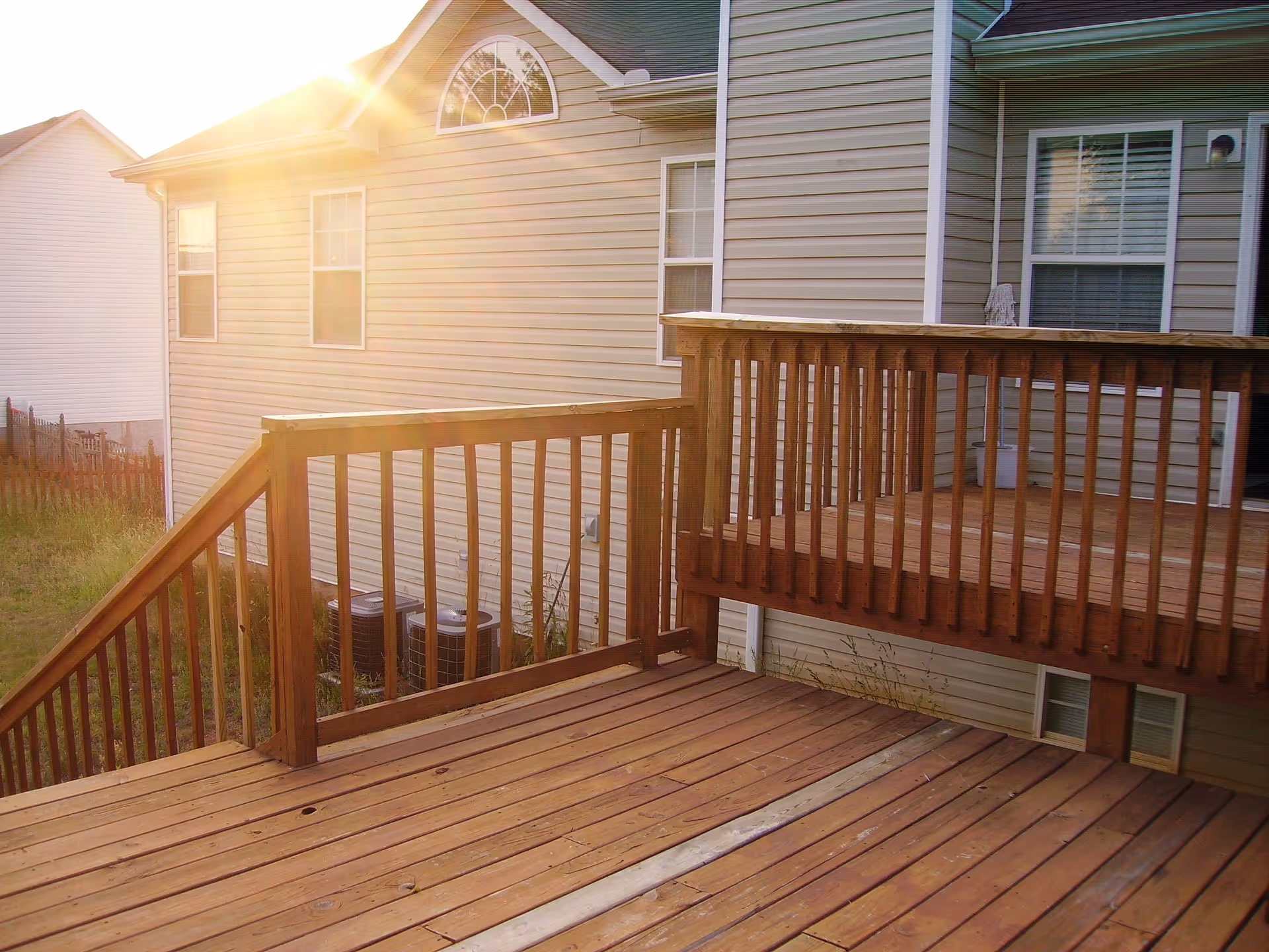 Sunlit wooden deck attached to the back of a beige house with white trim, featuring railings and stairs leading down to a grassy yard with neighboring houses visible.