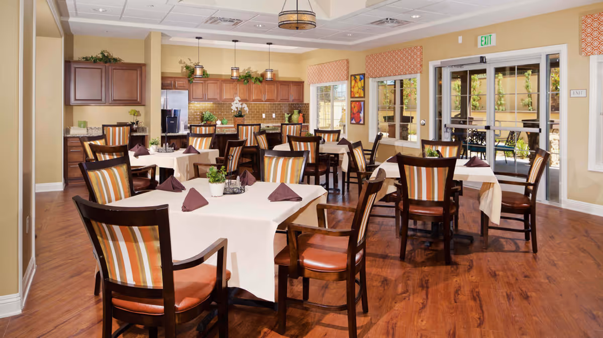 A dining room in a senior living facility with multiple square tables covered with beige tablecloths and brown napkins. Each table is surrounded by wooden chairs with striped upholstery. The room has wooden flooring, a kitchen area with wooden cabinets and a refrigerator in the background, and large windows with orange patterned valances. There is an exit door leading outside to a patio area with seating.