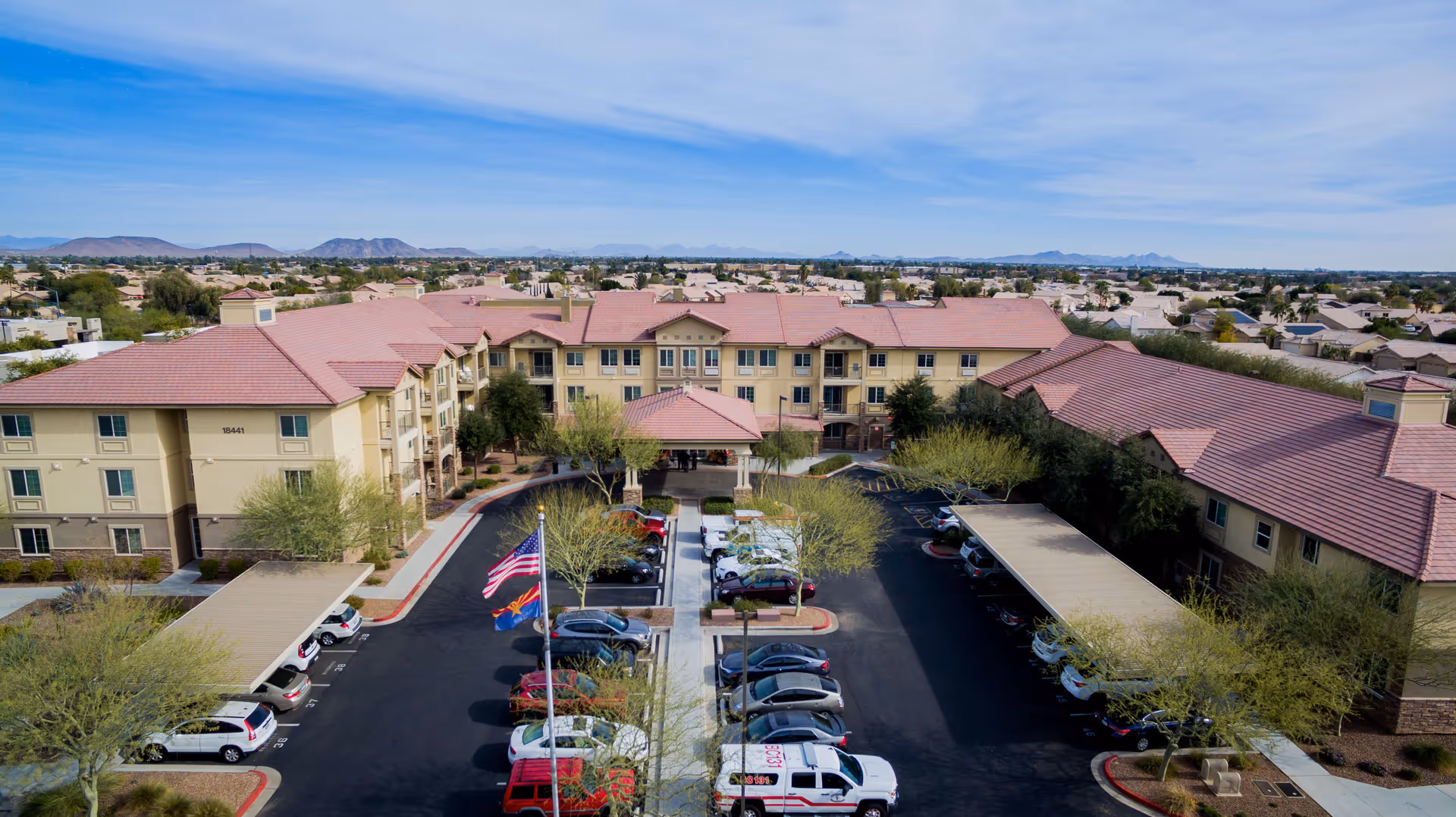 Aerial view of Palos Verdes Senior Living facility showing a large three-story building with a red-tiled roof surrounding a central parking lot with several cars parked. There are trees lining the parking area and a flagpole with American and state flags near the entrance. The background shows a suburban neighborhood and distant mountains under a partly cloudy sky.