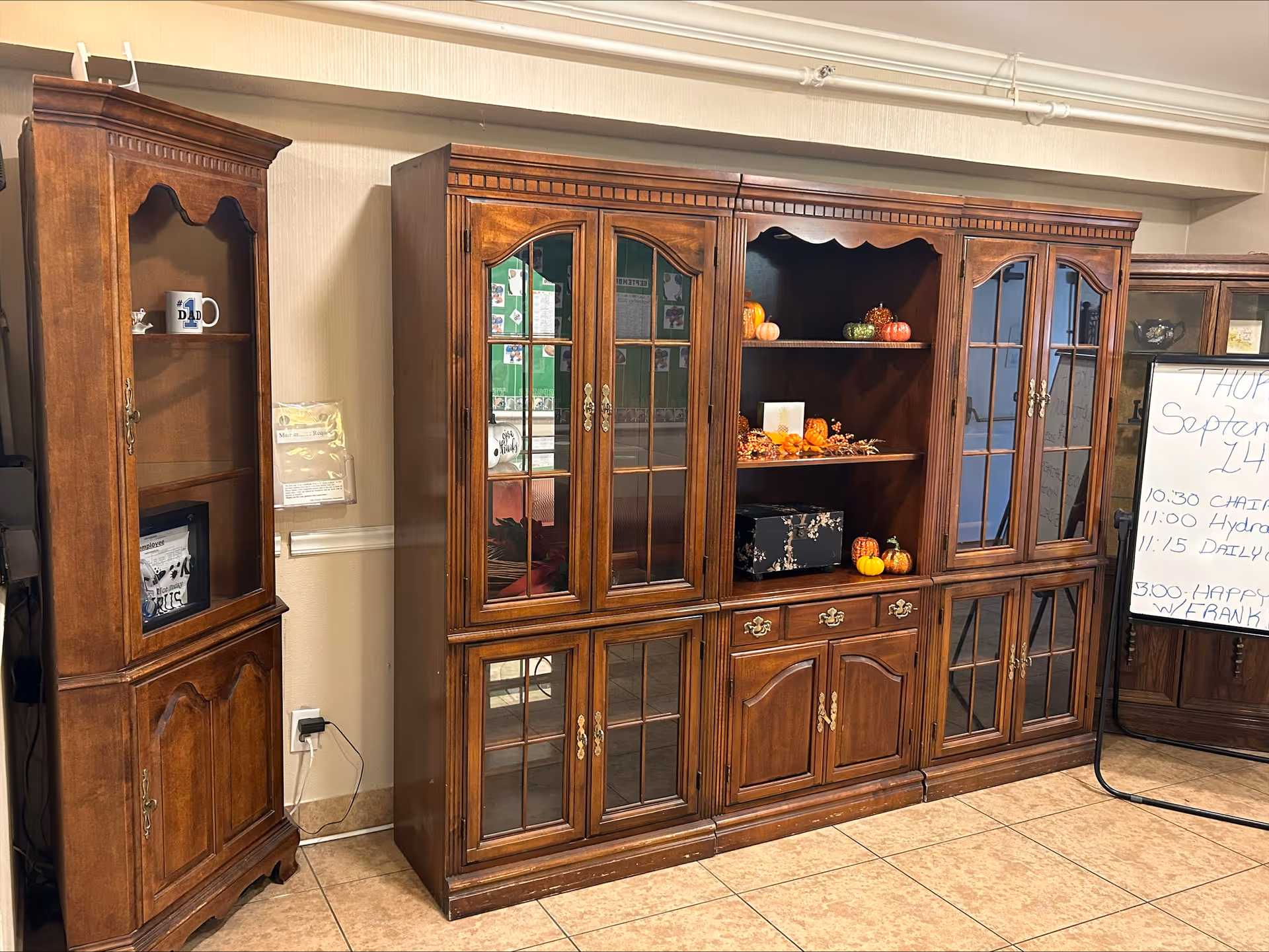 A room with wooden cabinets and shelves, some decorated with small pumpkins and fall-themed items. There is a whiteboard on the right side with a schedule written on it. The floor is tiled and the walls are light-colored.