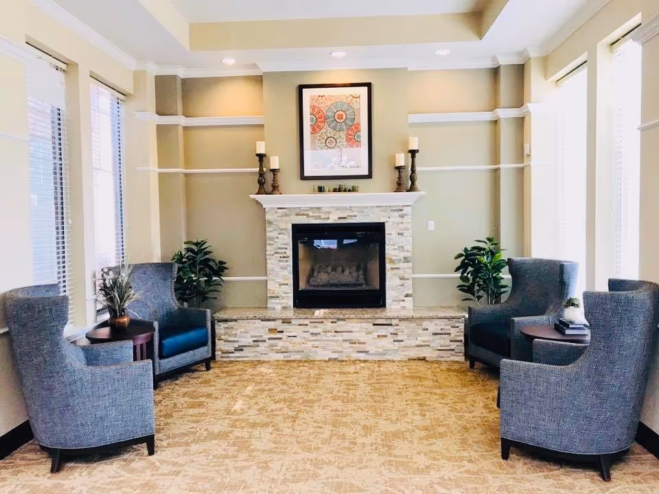 A cozy seating area with four gray upholstered armchairs arranged around a stone fireplace. The fireplace has a white mantel decorated with four candle holders and a framed abstract artwork above it. There are two small side tables with plants and books, and large windows with blinds on either side of the room, allowing natural light to fill the space.