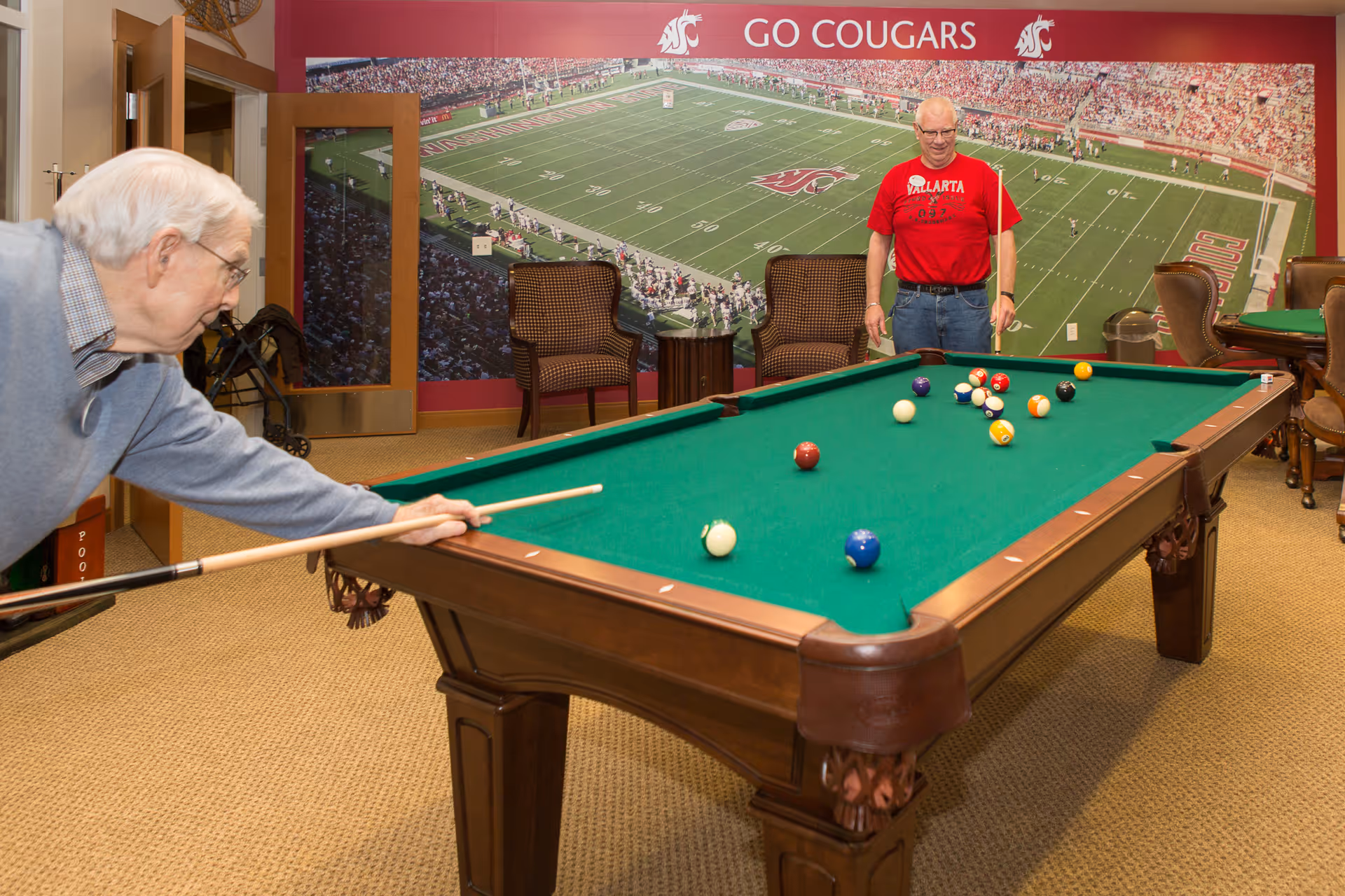 Two elderly men playing pool in a game room with a large mural of a football stadium and the words 'GO COUGARS' on the wall behind them. The room has carpeted flooring, chairs, and a door with glass panels.