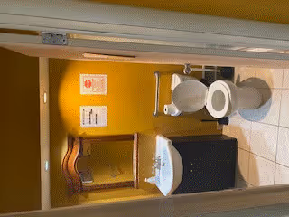 A small bathroom with a white toilet and a white sink with a dark cabinet underneath. Above the sink is a wooden framed mirror mounted on a yellow wall. There are two signs posted on the wall next to the mirror. The floor is tiled in white.