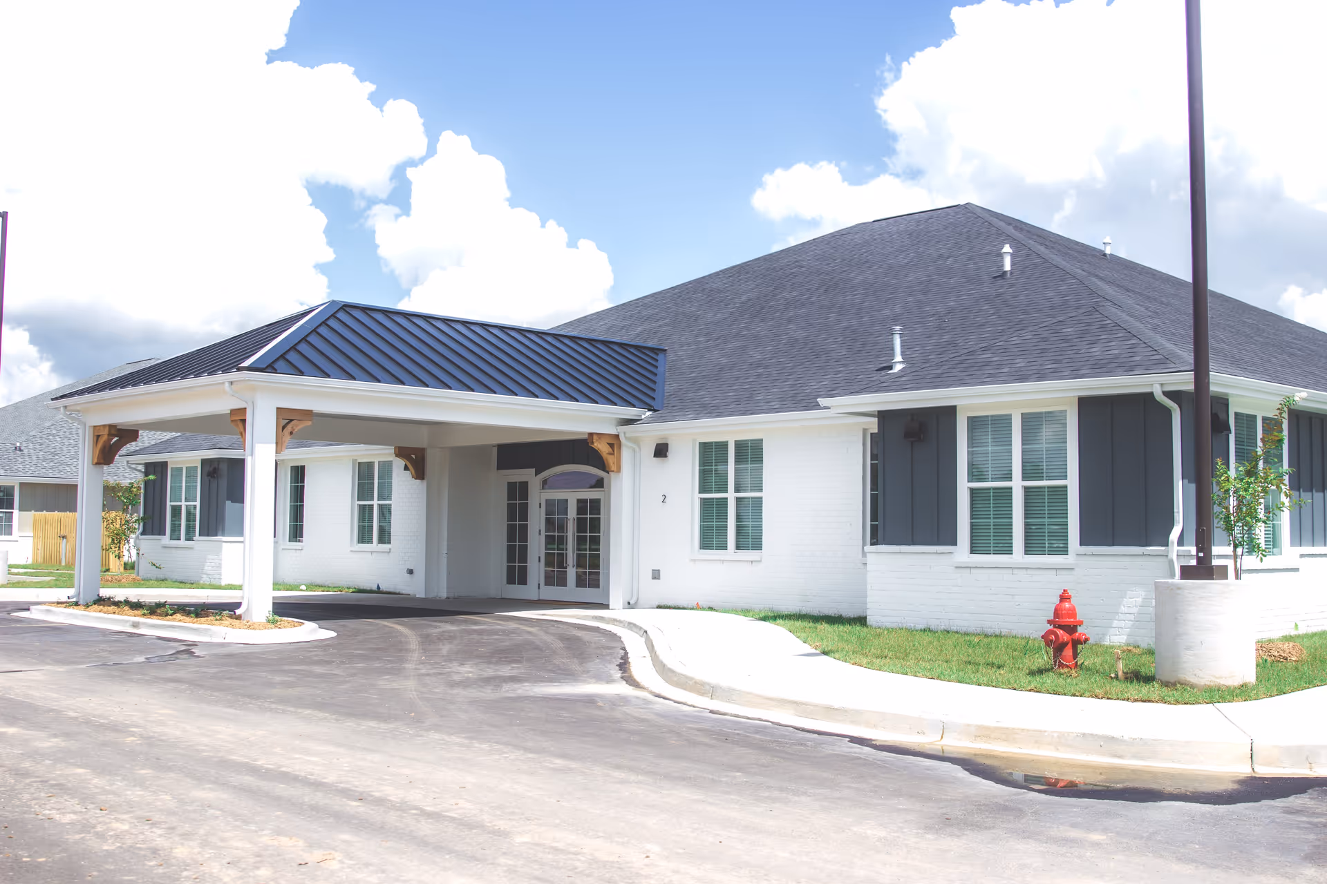 Exterior view of a single-story building with white brick walls and dark gray roof. The building has multiple windows with white frames and a covered entrance with a metal roof supported by white columns and wooden brackets. There is a red fire hydrant on the grass near the sidewalk and a streetlamp on the right side. The sky is partly cloudy with blue patches.