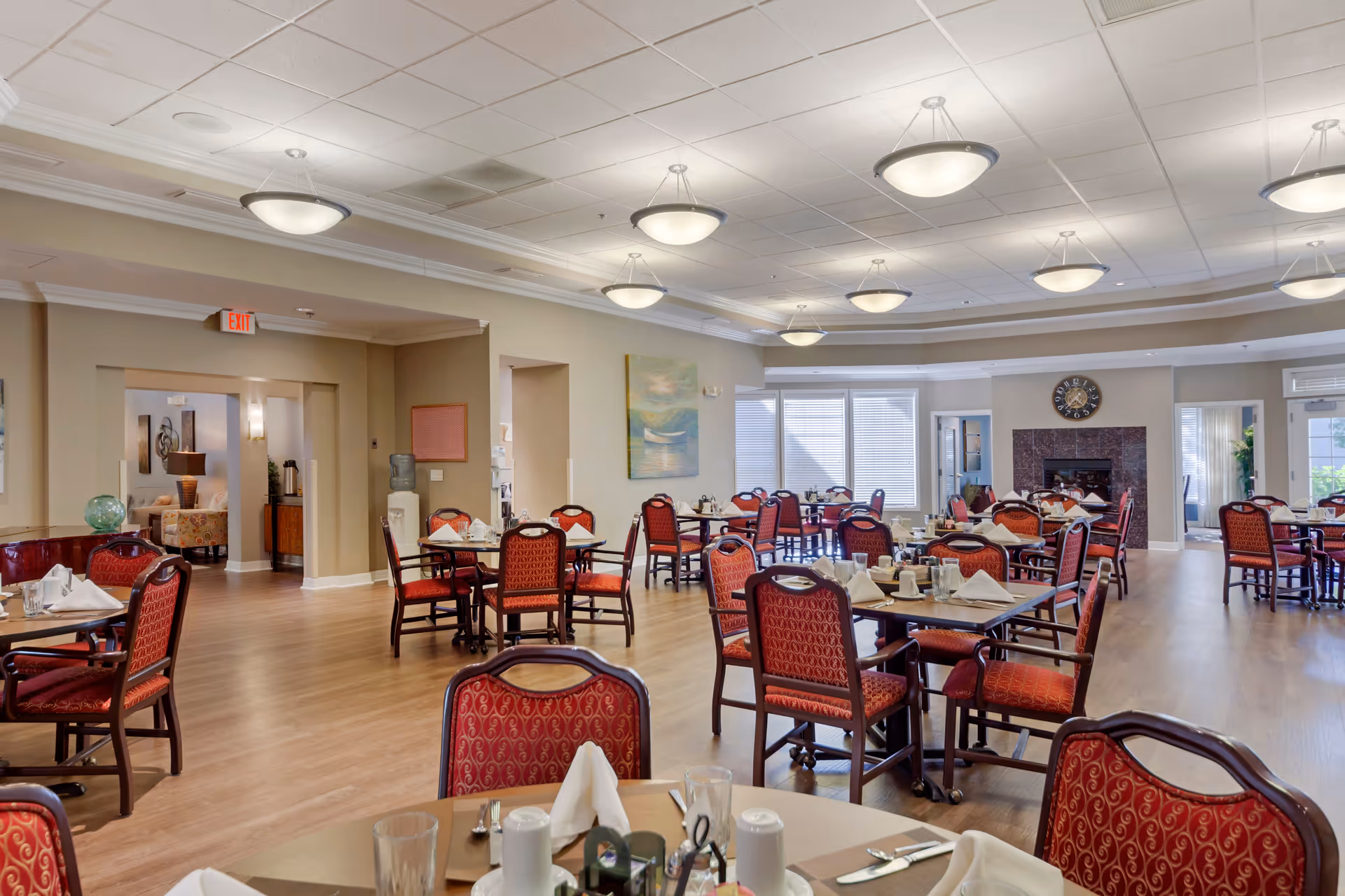 A spacious dining room in an assisted living facility with multiple round tables set with white napkins, glasses, and silverware. The room features red cushioned chairs with patterned upholstery, wooden flooring, and soft overhead lighting. Large windows with blinds allow natural light to fill the space. A fireplace with a clock above it is visible in the background, along with doorways leading to other rooms.