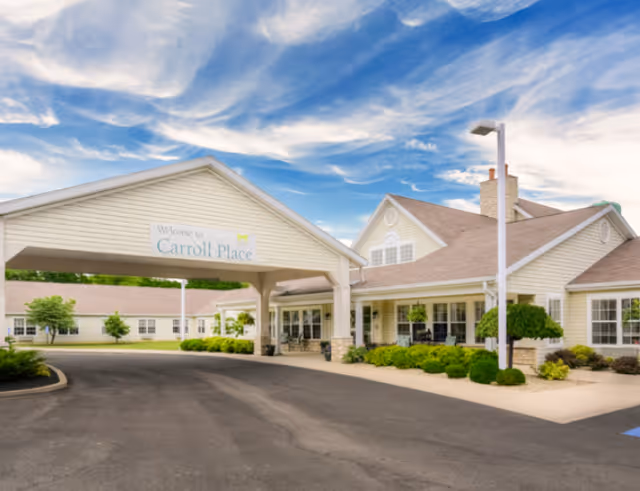 Exterior view of Carroll Place senior living facility with a covered entrance, beige siding, and a clear blue sky with some clouds.