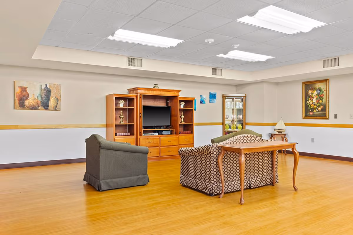 A senior living facility common area with wooden flooring, a patterned sofa, a gray armchair, a wooden entertainment center with a TV, and decorative paintings on the walls.