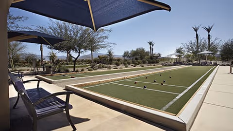Outdoor bocce ball court with benches and large blue umbrellas providing shade. Desert landscaping with trees and shrubs is visible in the background under a clear sky.