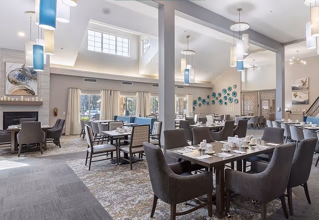 A spacious and well-lit dining room with multiple tables and chairs arranged neatly. The room features high ceilings with modern pendant lights hanging down, large windows with curtains allowing natural light to fill the space, and decorative wall art. The tables are set with white cups, plates, and napkins, ready for dining.