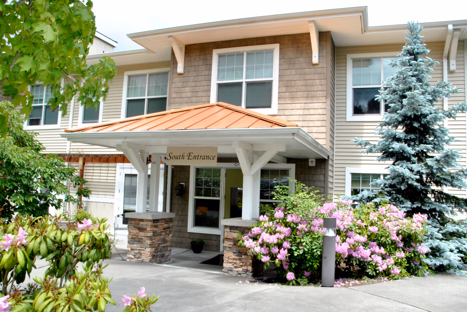 Exterior view of a senior living facility entrance labeled 'South Entrance' with a covered porch supported by white beams and stone pillars. The building has beige siding and multiple windows. There are green shrubs and pink flowers along the walkway, and a tall evergreen tree to the right.