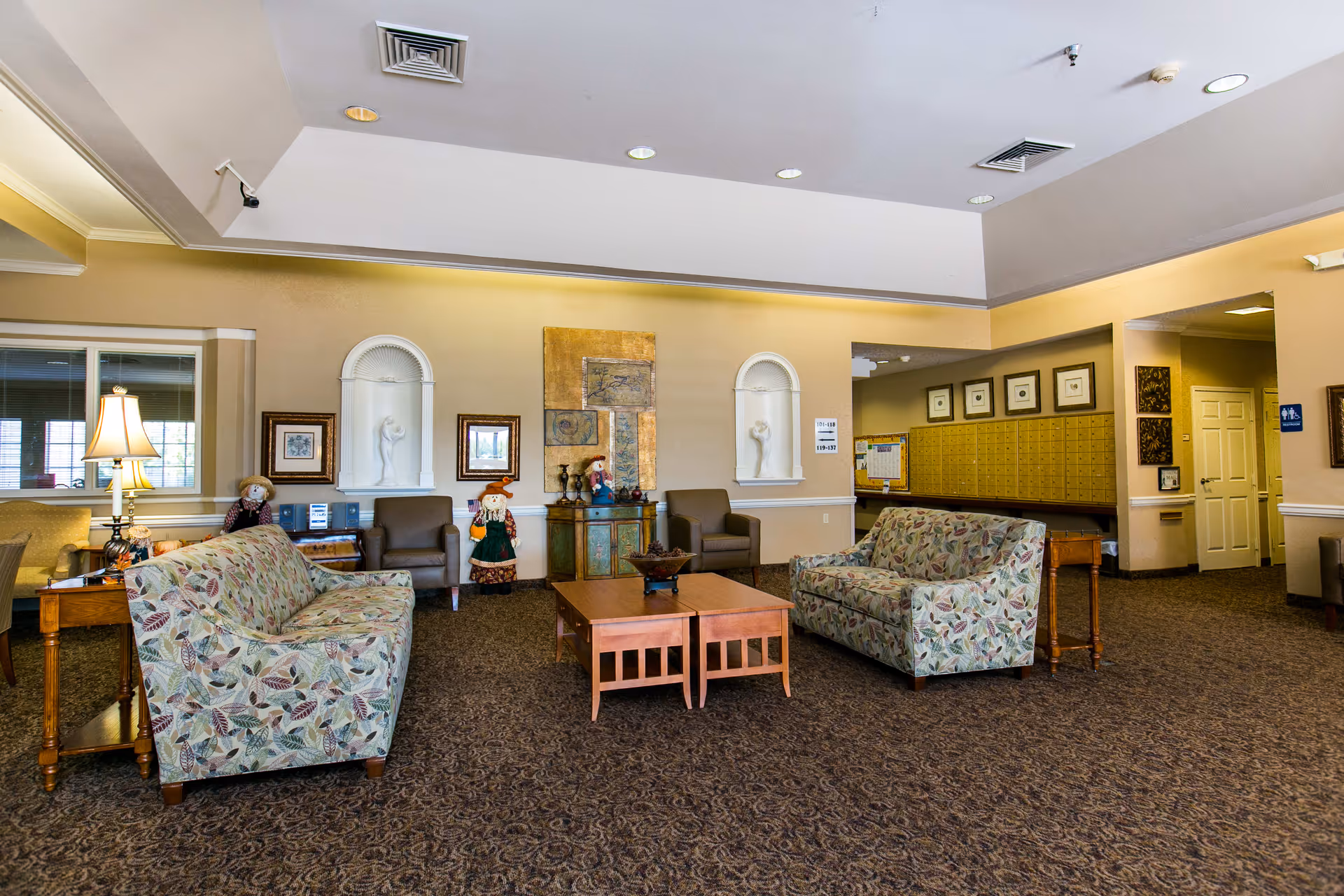 A spacious senior living facility common area with two floral patterned sofas facing each other and a wooden coffee table in the center. The room has beige walls with decorative niches containing white sculptures, framed artwork, and two scarecrow dolls. There are brown armchairs, a side table with a lamp, and a bulletin board with mailboxes in the background. The carpet is patterned and the ceiling has recessed lighting and ventilation.