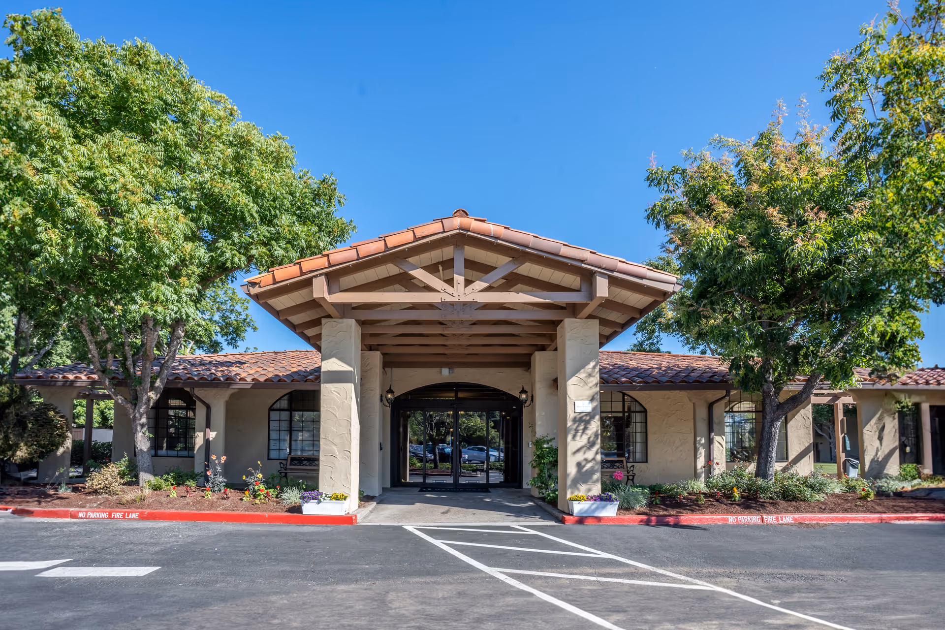 Front exterior view of a single-story building with a tiled roof and a covered entrance supported by columns. The building is surrounded by green trees and landscaped flower beds. The sky is clear and blue.