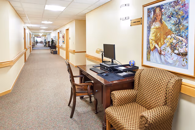 Well-lit interior corridor of a senior living facility with a desk, chairs, and artwork along the wall.