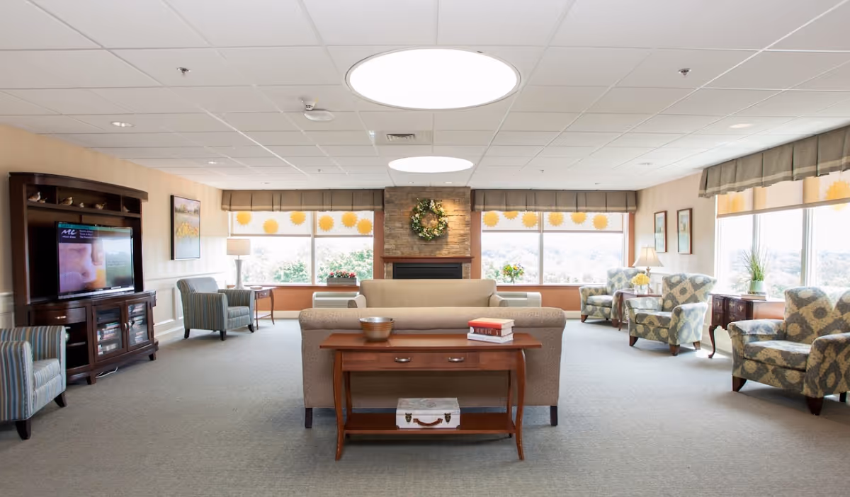 A spacious and well-lit living room area in a senior living facility with large windows letting in natural light. The room features a beige sofa facing a wooden console table with books and a decorative bowl. There are multiple patterned armchairs arranged around the room, a wooden entertainment center with a TV on the left, and a stone fireplace with a wreath above it in the center. The windows have valances and sun-shaped decals on the glass. The carpet is light gray, and the ceiling has recessed lighting and circular light fixtures.