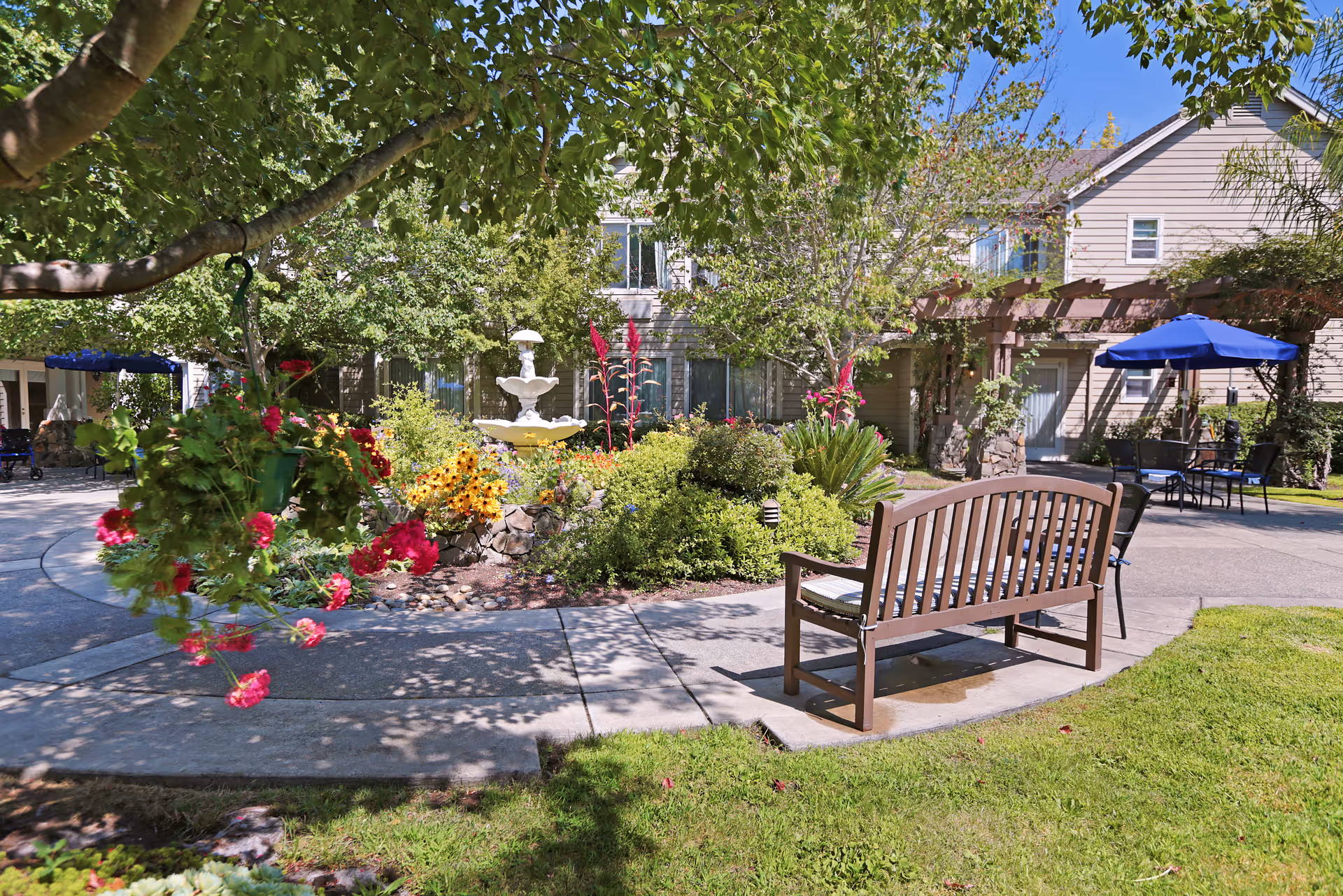 A peaceful outdoor garden area at a senior living facility with a wooden bench, hanging flower baskets, colorful flower beds, a white tiered fountain, and patio tables with blue umbrellas. Trees provide shade and a building with windows and a pergola is visible in the background.