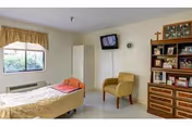 A private senior care bedroom with a single bed, wall-mounted TV, armchair, window, and a shelving unit.