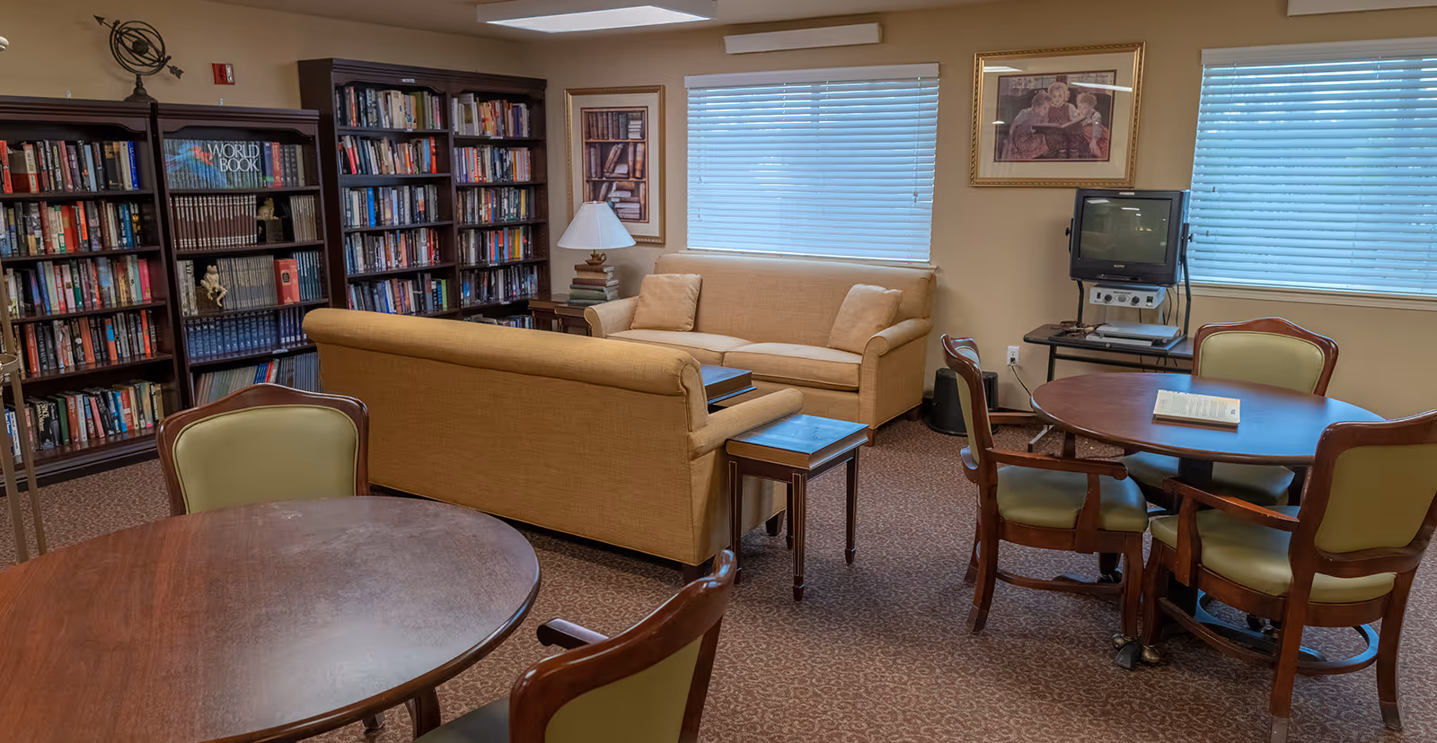 Cozy communal living area with bookshelves, couches, round tables and chairs, and a small TV in a senior living facility.