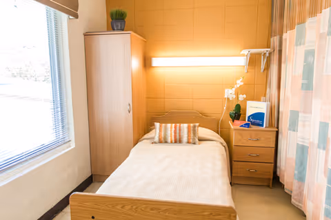 A small, simple bedroom in a care facility featuring a single bed with a beige bedspread and a decorative pillow. Next to the bed is a wooden nightstand with a white orchid plant, a lamp, and some brochures. A wooden wardrobe stands against the wall near a window with blinds, and a curtain with a pastel geometric pattern hangs on the right side. The walls are painted a warm yellow color.