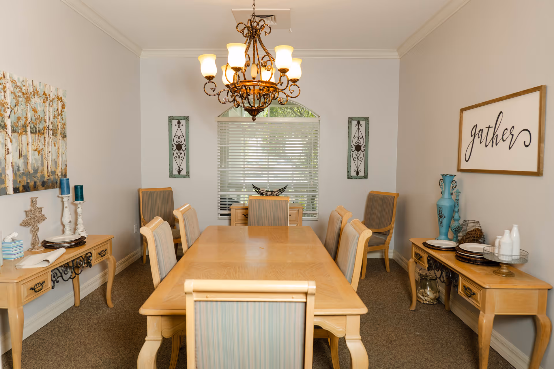 A dining room with a wooden table surrounded by eight chairs with light upholstery. The room features a decorative chandelier hanging above the table, a window with blinds at the far end, and two side tables against the walls. One side table has candles, plates, and decorative items, while the other has a large blue vase, plates, and a sign on the wall that says 'gather'. The walls are painted light gray and the floor is carpeted.