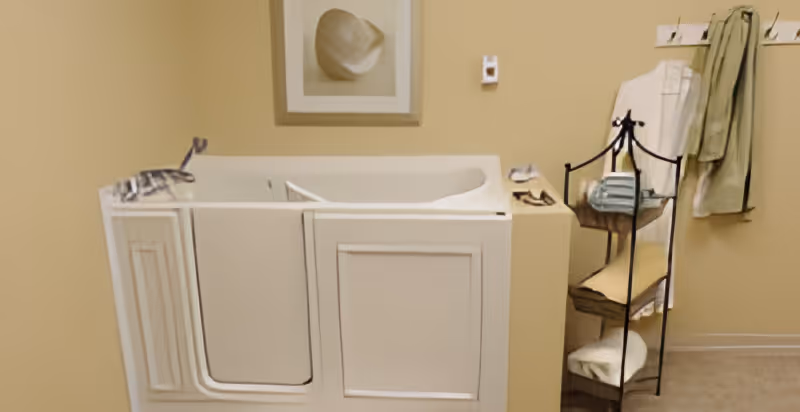 Interior view of a bathroom featuring a walk-in bathtub with a door, a towel rack with folded towels, a wall-mounted hook with hanging towels, and a framed picture of a hat on the wall.
