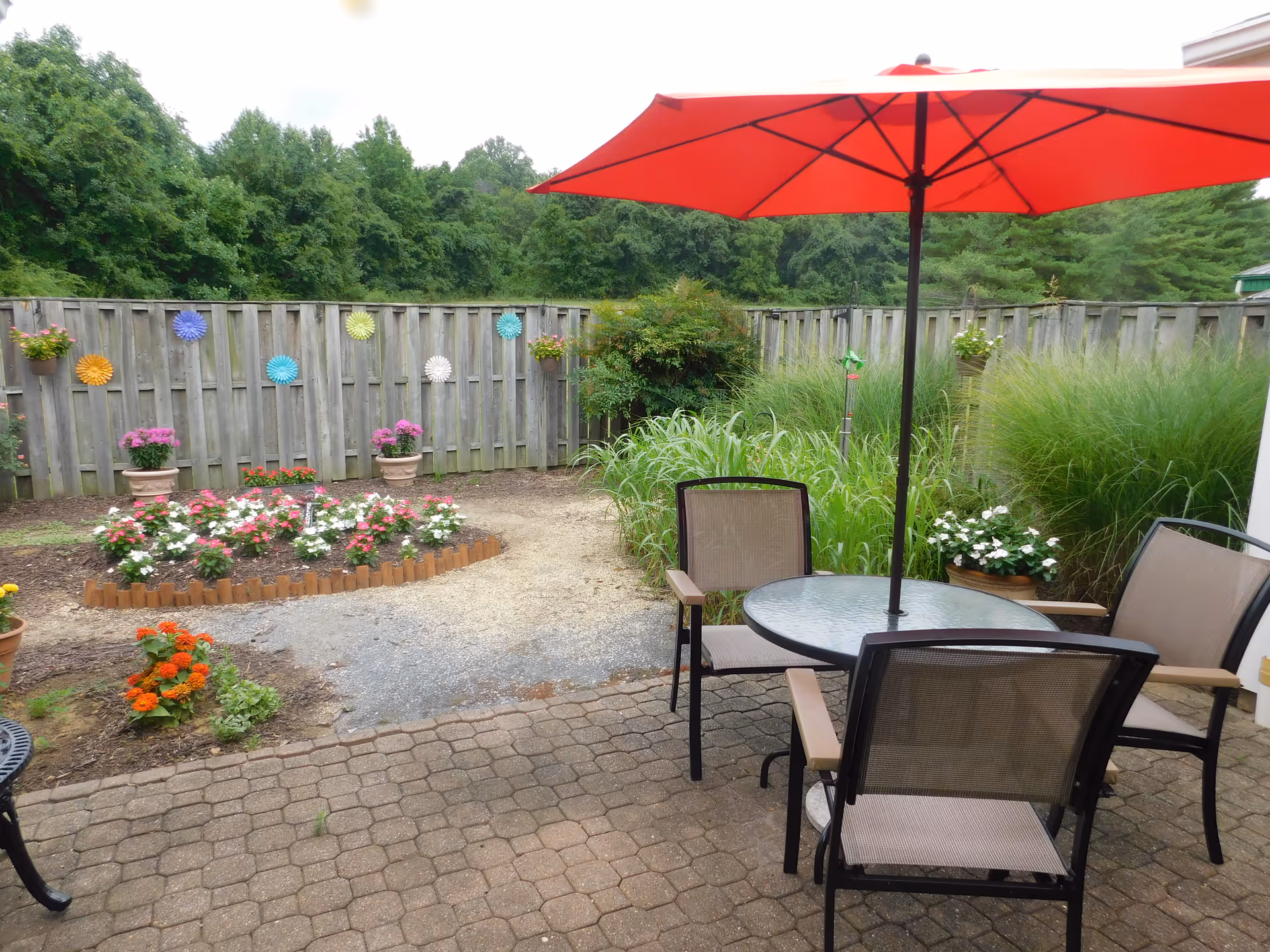Patio area with a round glass table, chairs and a red umbrella next to flower beds and a wooden fence decorated with colorful ornaments.