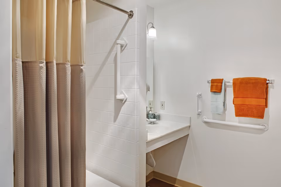 A clean bathroom with a beige and brown shower curtain, white tiled shower area with a grab bar, a white sink with a mirror above it, and a wall-mounted towel rack holding orange and light blue towels. The bathroom has white walls and a light fixture above the mirror.