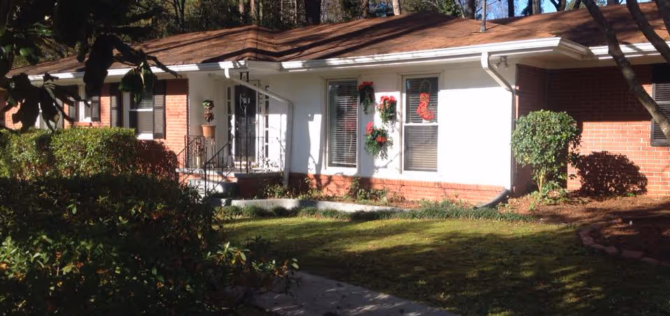 Exterior view of a single-story brick and white siding building with a brown roof, surrounded by bushes and trees. The entrance has a small set of stairs with a black railing, and the windows are decorated with wreaths and holiday decorations.